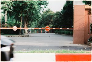 A stop barrier closes off a city street entrance bordered by green trees.